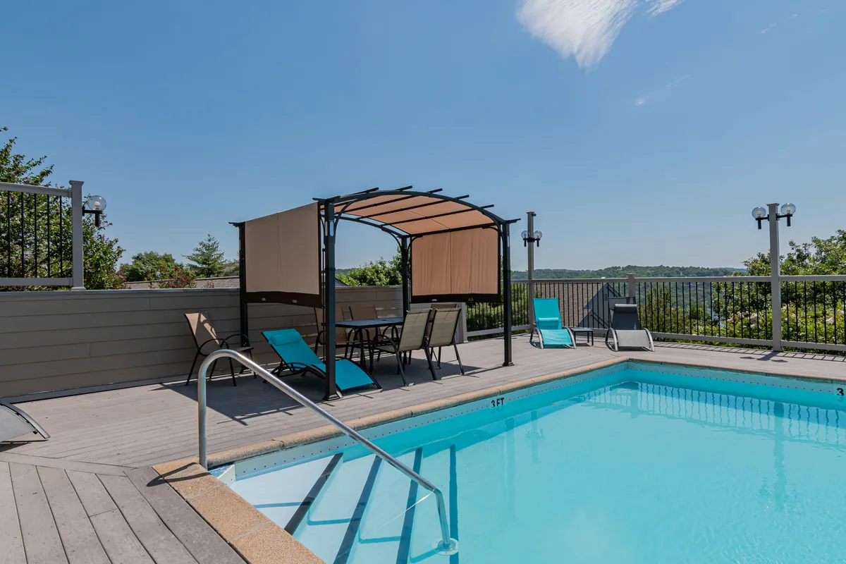 A vibrant outdoor swimming pool area featuring a sparkling pool surrounded by lounge chairs and a shaded canopy seating area, all set against a scenic landscape backdrop. The space is designed for leisure, socializing, and enjoying sunny days with friends or family.