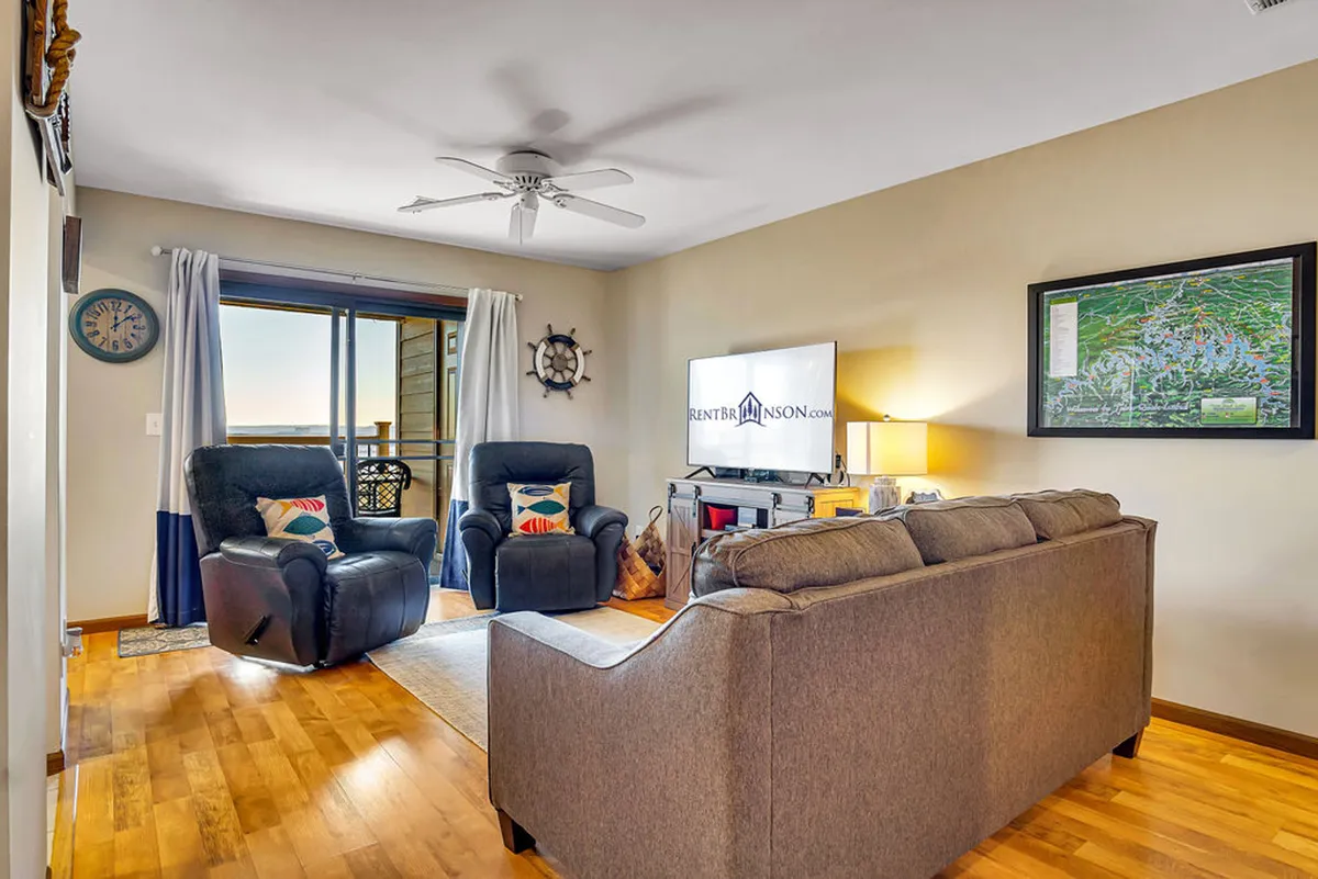 This image features a cozy living room with comfortable recliners, a prominent television, and a large window that frames a scenic view. The space is sunlit, creating a welcoming atmosphere suitable for relaxing or gathering with loved ones.