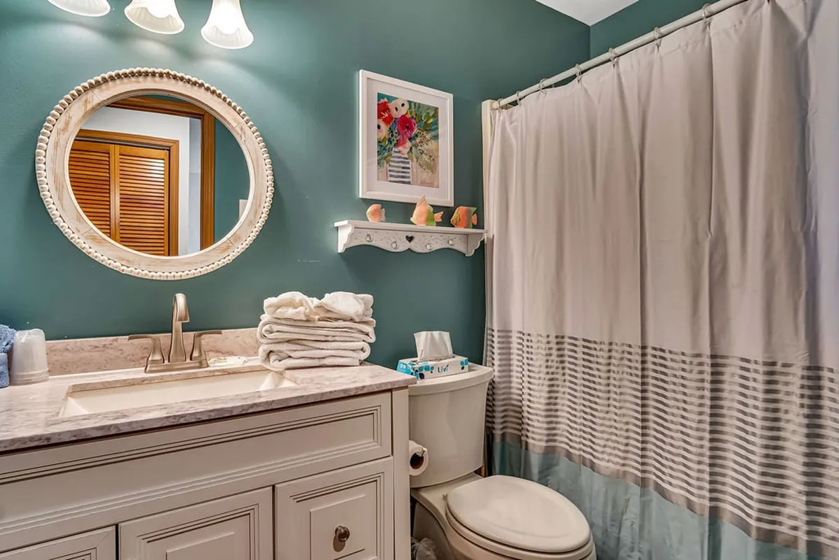 A fresh and cheerful bathroom featuring a circular mirror above the vanity, a modern sink, decorative artwork, and a striped shower curtain. The space feels inviting and well-kept, ideal for morning or evening routines.