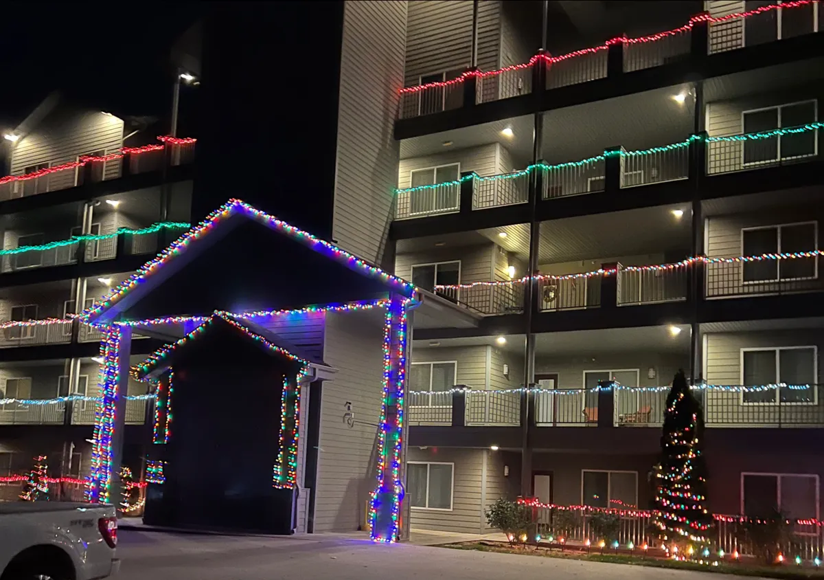A festive exterior view of the property, adorned with Christmas lights on balconies, a decorated entrance, and a Christmas tree out front. The scene radiates warmth and a welcoming spirit, hinting at shared holiday memories and social gatherings.