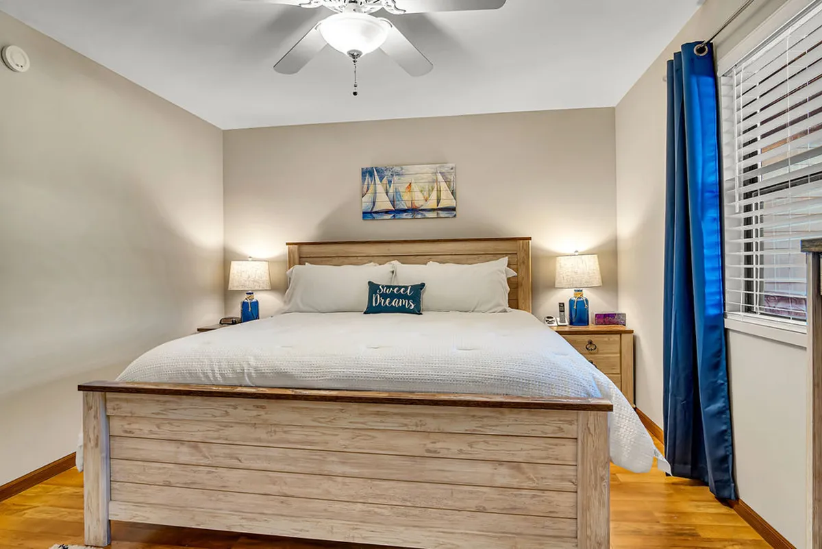 A cozy bedroom with a king-size bed dressed in inviting linens, decorative pillows, matching nightstands, and tasteful wall art. Soft light filters in through the curtains, creating a tranquil atmosphere ideal for rest and relaxation.