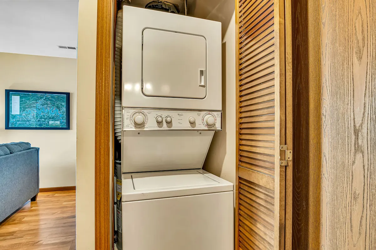 A practical laundry area with a stackable washer and dryer set neatly behind wooden louvered doors, complemented by a piece of framed artwork in the background. The space is tidy and functional, ideal for hassle-free laundry during longer stays.